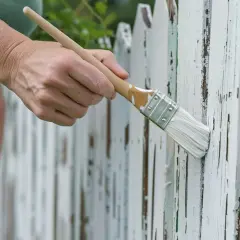 Ręka z pędzlem odnawia biały, drewniany płot. Stara farba łuszczy się, ukazując surowe drewno.