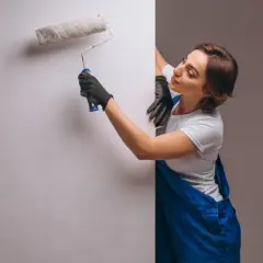 Kobieta w niebieskim ogrodniczkach i czarnych rękawiczkach maluje ścianę wałkiem.