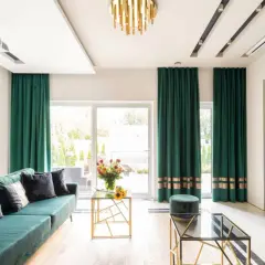 Elegancki salon z zielonymi zasłonami przy oknach. Wygodna kanapa, szklane stoliki i nowoczesny telewizor tworzą przytulną atmosferę.