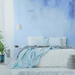 Sypialnia z łóżkiem i niebieskim ombre na ścianie. Delikatne światło i rośliny tworzą spokojną atmosferę.
