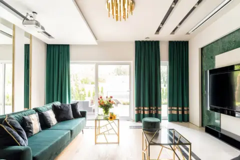 Elegancki salon z zielonymi zasłonami przy oknach. Wygodna kanapa, szklane stoliki i nowoczesny telewizor tworzą przytulną atmosferę.