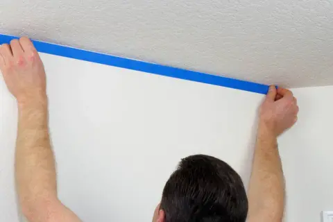 Ręce przykładają niebieską taśmę malarską, tworząc odcięcie sufitu od ściany.