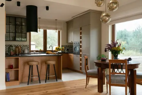 Nowoczesna kuchnia w stylu modern farmhouse z drewnianą wyspą, szarymi frontami i dużym oknem z widokiem na las.