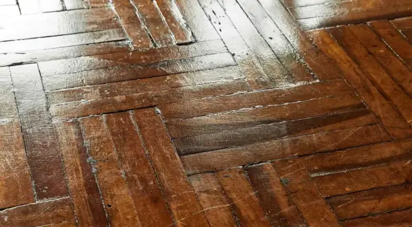 Wytarty lakier na parkiecie, widoczne ślady użytkowania i zmatowienia na drewnianej podłodze.