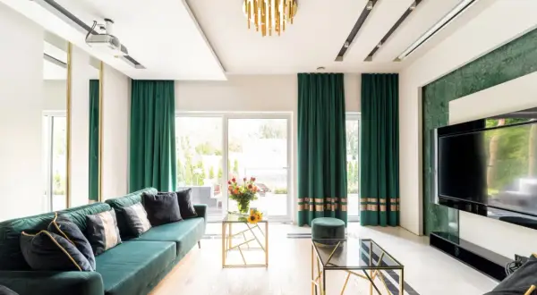Elegancki salon z zielonymi zasłonami przy oknach. Wygodna kanapa, szklane stoliki i nowoczesny telewizor tworzą przytulną atmosferę.