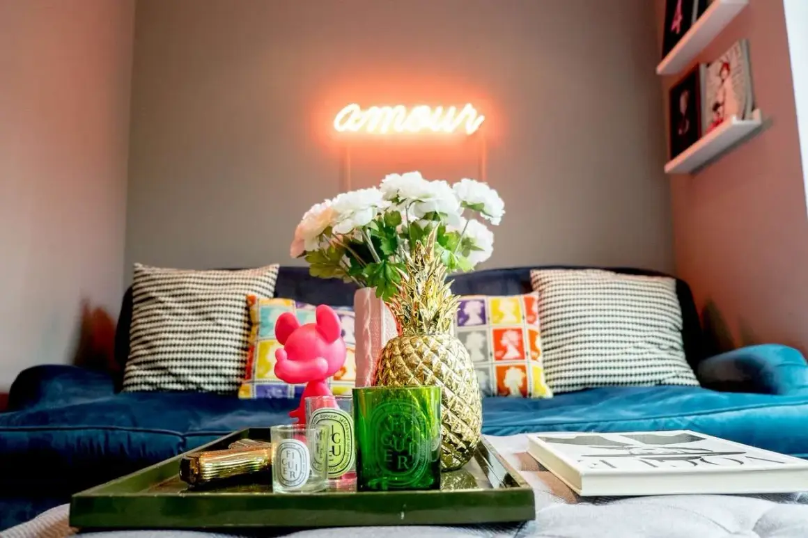 Jak ocieplić mieszkanie dodatkami: neon "amour", złoty ananas, świece i kwiaty na granatowej kanapie tworzą przytulną atmosferę.