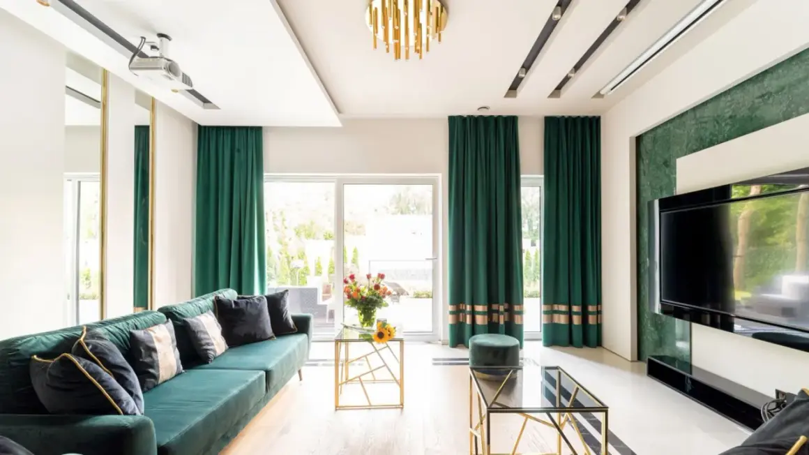 Elegancki salon z zielonymi zasłonami przy oknach. Wygodna kanapa, szklane stoliki i nowoczesny telewizor tworzą przytulną atmosferę.