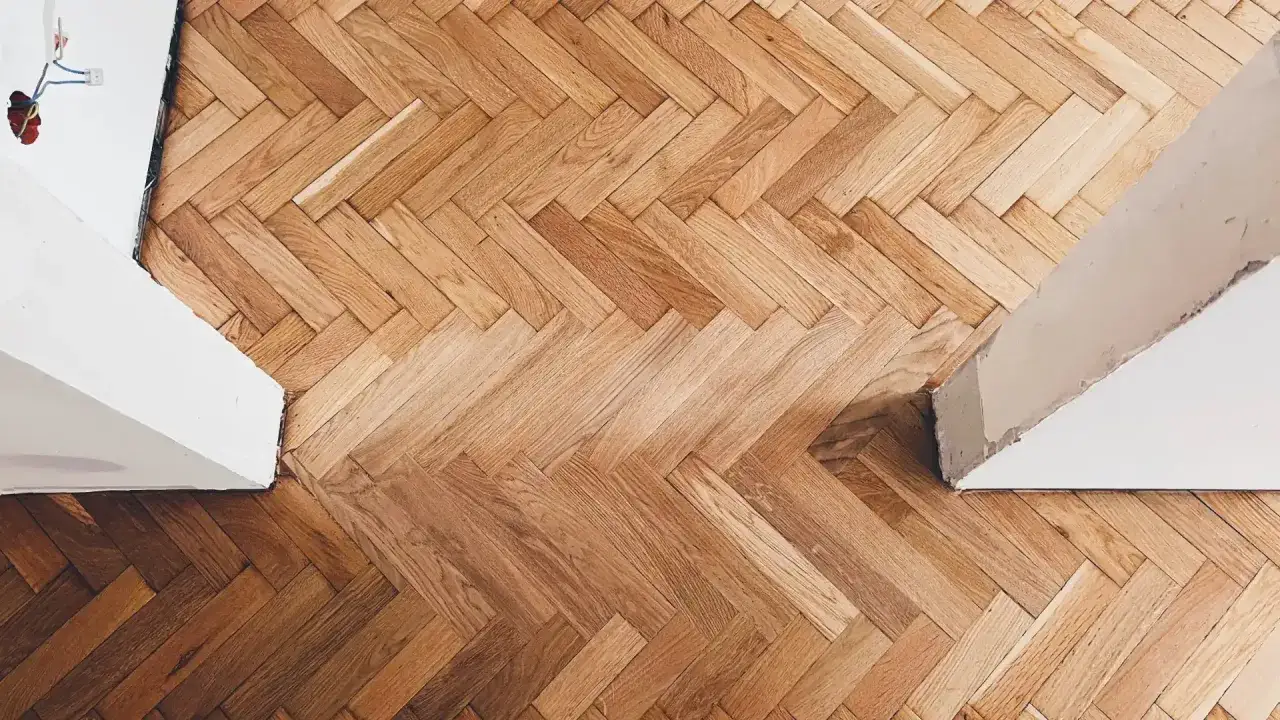 Drewniany parkiet ułożony we wz&oacute;r jodełki, z widocznym wytartym lakierem w miejscach intensywnego użytkowania.