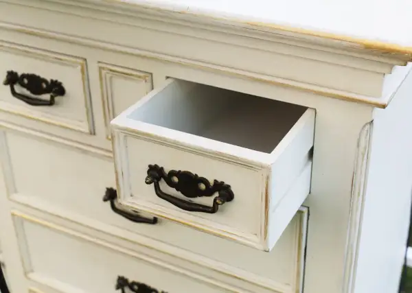 Biała komoda z ozdobnymi uchwytami, jedna szuflada lekko wysunięta. Inspiracje na odnawianie starych mebli.