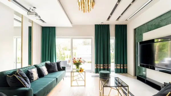 Elegancki salon z zielonymi zasłonami przy oknach. Wygodna kanapa, szklane stoliki i nowoczesny telewizor tworzą przytulną atmosferę.