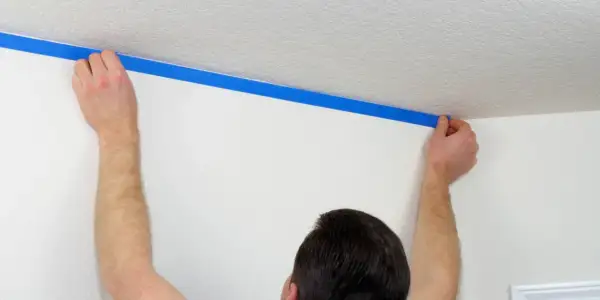 Ręce przykładają niebieską taśmę malarską, tworząc odcięcie sufitu od ściany.